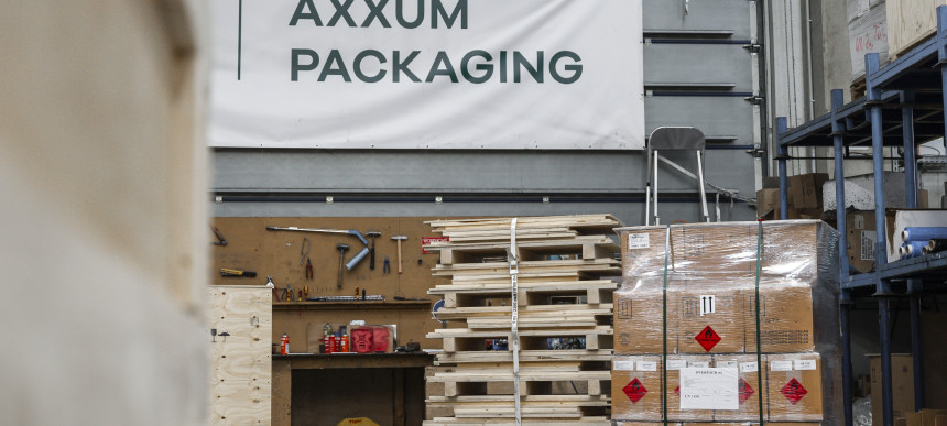 Lagerhalle mit gestapelten Holzpaletten, verpackten Kartons auf einer Palette und einem Schild mit der Aufschrift AXXUM PACKAGING