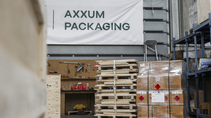 Lagerhalle mit gestapelten Holzpaletten, verpackten Kartons auf einer Palette und einem Schild mit der Aufschrift AXXUM PACKAGING