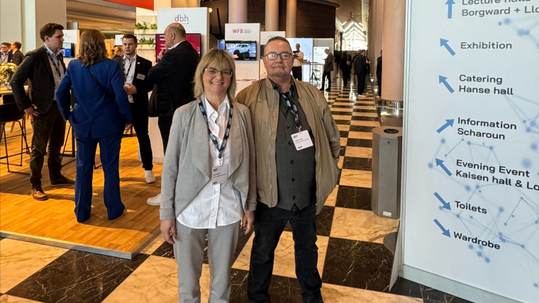 Zwei Personen stehen in einer Halle mit schwarz-weiß kariertem Boden vor einem Wegweiser der Veranstaltung Logistics Connect