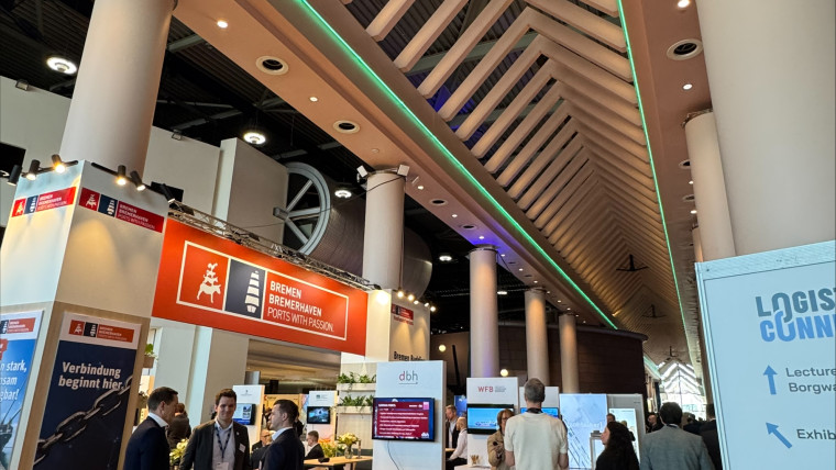 Messestand mit mehreren Personen in einem großen, modernen Ausstellungsraum mit hohen Säulen und sichtbarer Dachkonstruktion