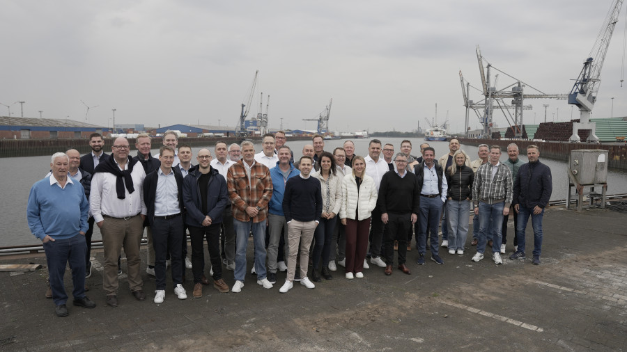 Eine Gruppe von Personen in Freizeitkleidung steht auf einem Industriedock mit Kränen und Containern im Hintergrund unter einem bewölkten Himmel; sie wirken fröhlich.