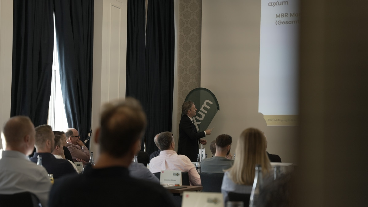 Ein Redner hält vor einem aufmerksamen Publikum in einem schwach beleuchteten Konferenzraum eine Präsentation, wobei auf einer Projektionsfläche eine Präsentation und das „AXXUM“-Branding zu sehen sind.