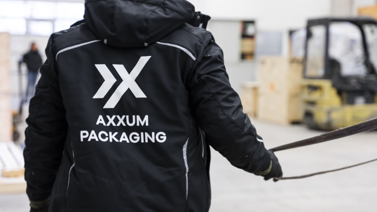 Eine Person in einer schwarzen Jacke mit der Aufschrift „Axxum Packaging” auf dem Rücken zieht in einem Lagerhaus an einem Gurt. Im Hintergrund sind ein Gabelstapler und Holzkisten zu sehen.