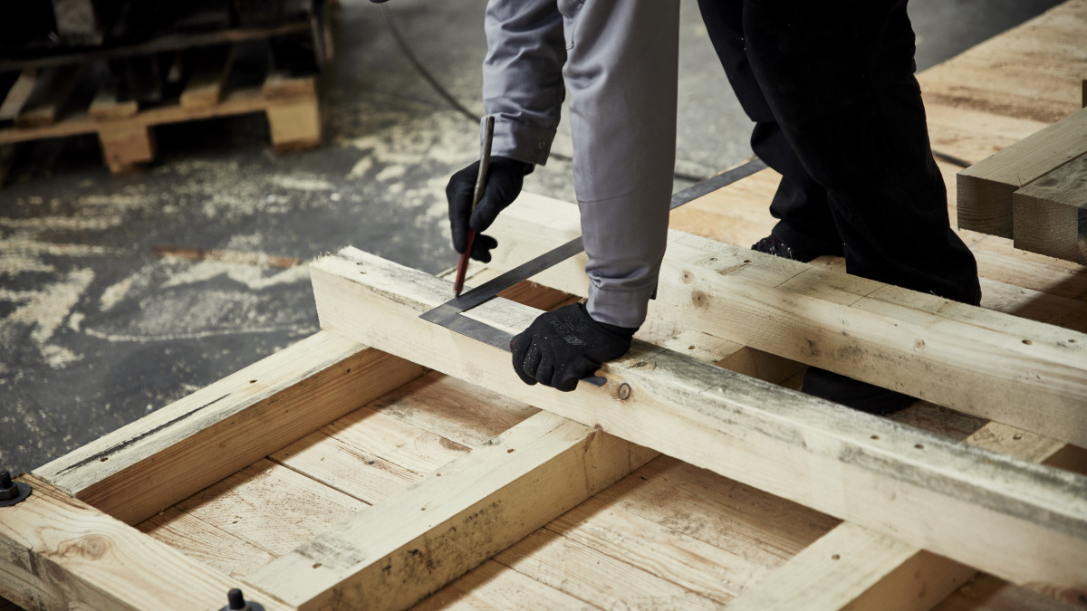 Eine Person, die Handschuhe und Arbeitskleidung trägt, misst in einer Werkstatt mit einem Winkelmaß Holzplanken.