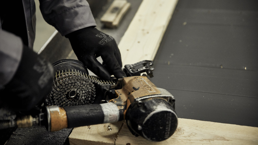 Eine Person mit schwarzen Handschuhen befestigt mit einer Druckluftnagelpistole Holzplanken aneinander.