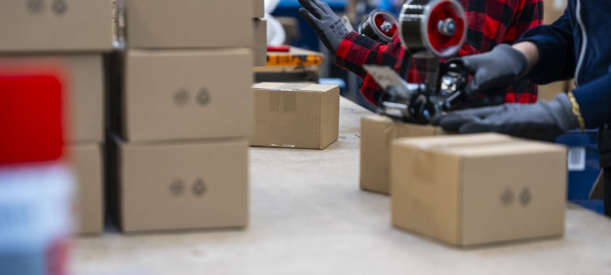 Arbeiter in einem Lagerhaus packen Kartons auf einem Tisch. Einer trägt ein rotes kariertes Hemd, der andere benutzt einen Klebebandspender.
