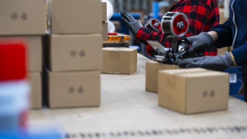 Arbeiter in einem Lagerhaus packen Kartons auf einem Tisch. Einer trägt ein rotes kariertes Hemd, der andere benutzt einen Klebebandspender.