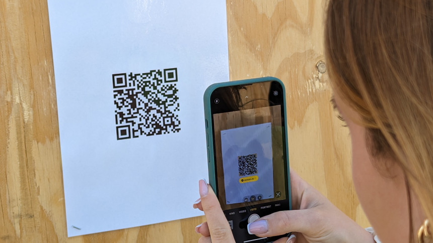 Frau hält Smartphone und scannt QR-Code auf weißem Blatt an Holzplatte