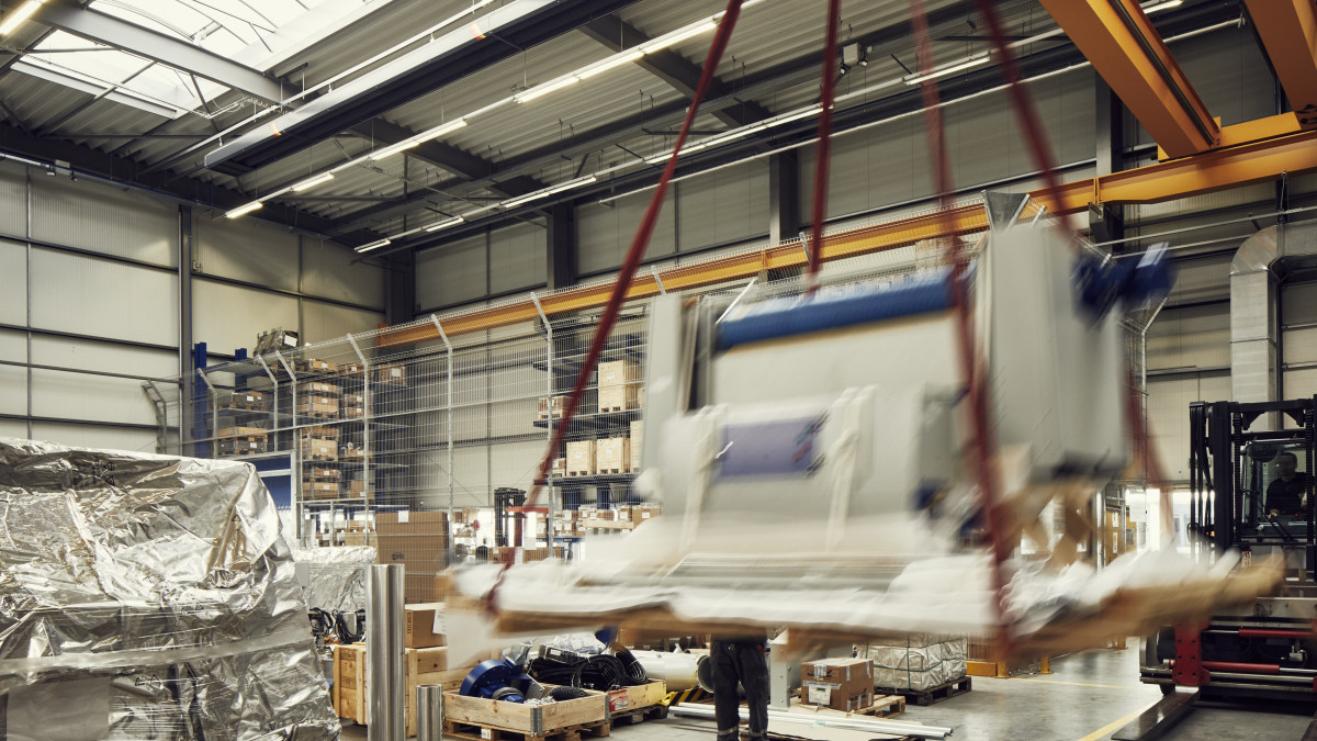 Eine Lagerhalle, wo Maschinen montiert werden. Man sieht verschiedene Maschinenteile. Im Vordergrund wird gerade ein Teil mit einem Kran angehoben.