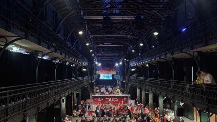Blick in eine große Halle mit hoher Decke, zwei weißen Fahrzeugen im Vordergrund und vielen Menschen, die sich in der Mitte versammeln