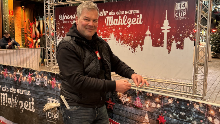 Mann in schwarzer Jacke und Jeans steht vor weihnachtlich dekoriertem Banner mit Schriftzug Mehr als eine warme Mahlzeit in einer Halle mit Gitterbalken und Glasdach