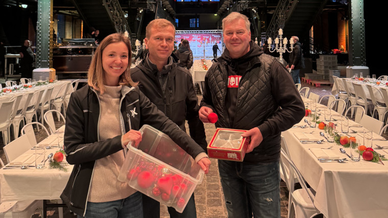 Drei Personen in warmer Kleidung stehen in einem festlich gedeckten, langgestreckten Saal, eine Person hält eine durchsichtige Box mit roten Bällen, eine andere eine rote Schachtel, im Hintergrund hängt ein Banner mit der Aufschrift Mehr als eine warme Mahlzeit