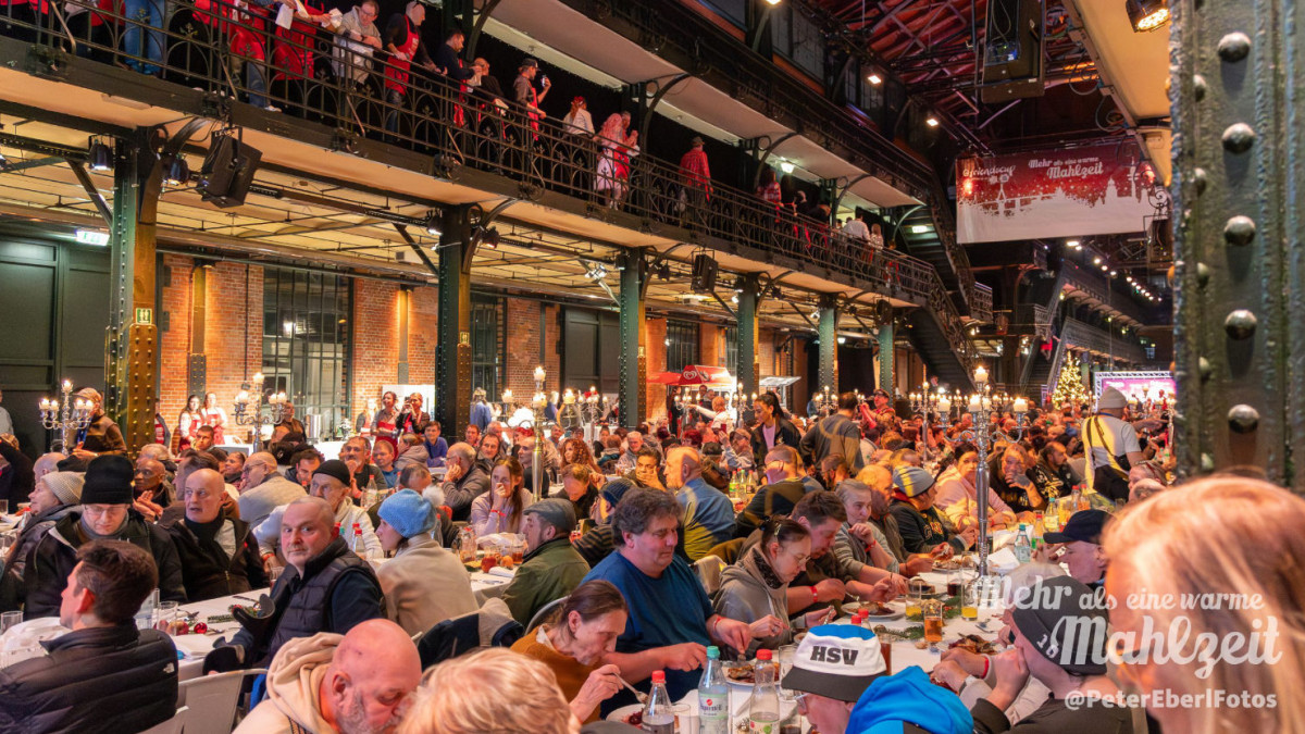 Große Menschenmenge sitzt an langen Tischen in einer Halle mit Stahlträgern und Galerie, viele essen und unterhalten sich