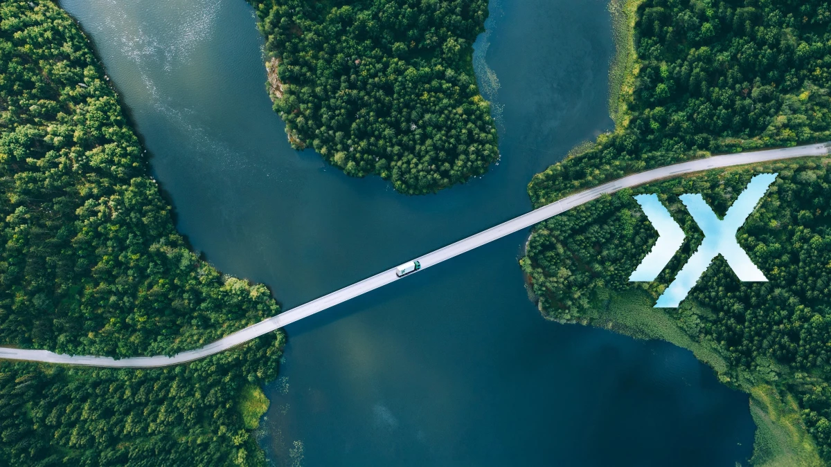 Luftaufnahme einer schmalen Brücke über einen Fluss, umgeben von üppig grünen Wäldern. Ein einzelnes Auto überquert die Brücke und vermittelt einen Eindruck von ruhiger Abgeschiedenheit. Auf der rechten Seite ist ein „XX“ zu sehen.