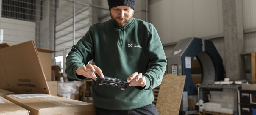 Ein Mann in einem grünen Pullover und einer schwarzen Mütze scannt ein Paket mit einem Handgerät in einem Lagerhaus, umgeben von Kartons und Industrieanlagen.