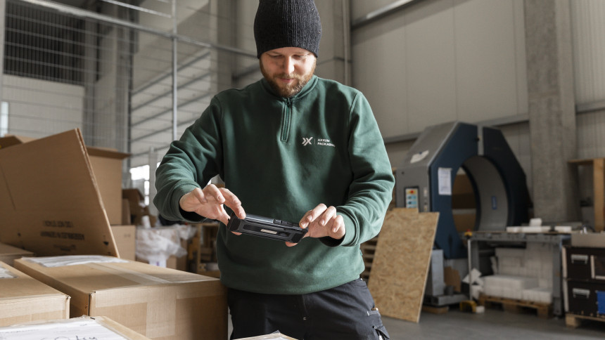 Ein Mann in einem grünen Pullover und einer schwarzen Mütze scannt ein Paket mit einem Handgerät in einem Lagerhaus, umgeben von Kartons und Industrieanlagen.