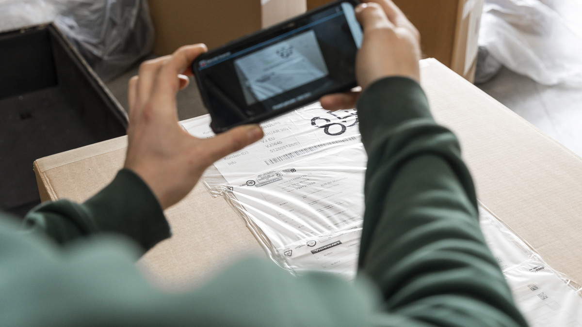 Eine Person in einem grünen Pullover scannt mit einem Smartphone einen Barcode auf einem großen Karton in einem Lagerhaus, umgeben von anderen Kartons.