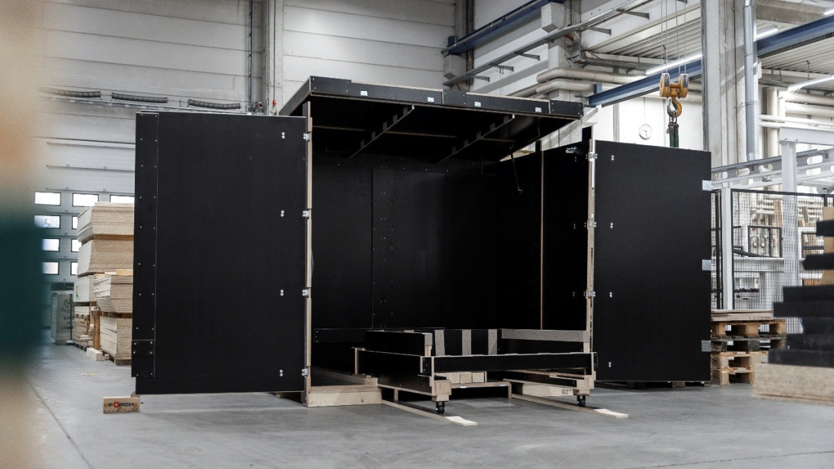 Eine große schwarze Transportkiste, teilweise zusammengebaut, steht in einem Industrielager mit sichtbaren Paletten und Rohren.