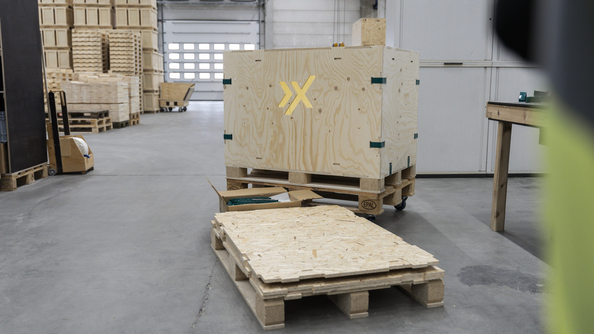 AXXUM kliXXkiste mit gelbem X-Symbol auf Holzpalette in Lagerhalle mit weiteren gestapelten Holzkisten
