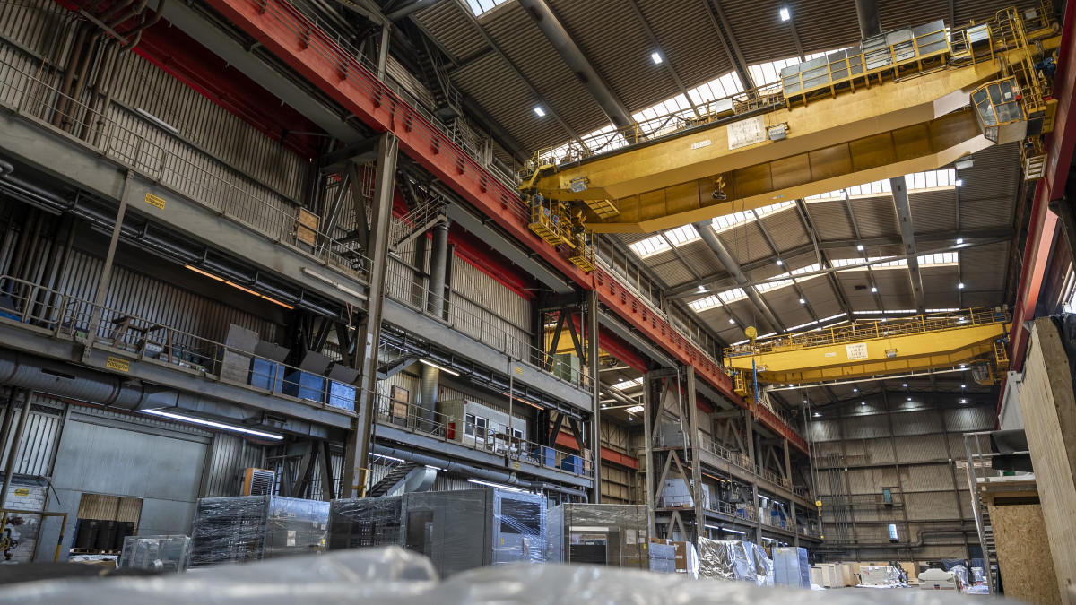 Innenraum eines großen Industrielagers mit Brückenkränen, Metallkonstruktionen, Regalen und verschiedenen Paletten und Geräten auf dem Boden.